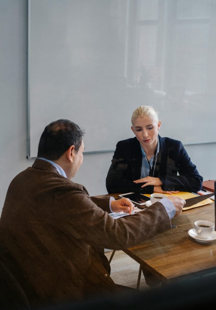 Smart colleagues in formal outfit discussing business while sitting at table with cups of coffee in office with whiteboard